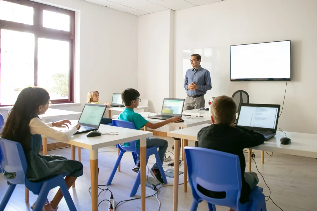 A modern classroom where students are using laptops during a lesson with a teacher instructing at the front, illustrating Smart Classroom Solutions in Kerala.